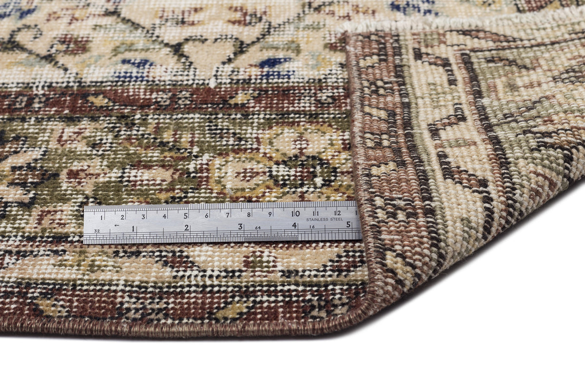 Beige, brown, and olive green traditional floral rug with a folded corner and a metal ruler for scale.