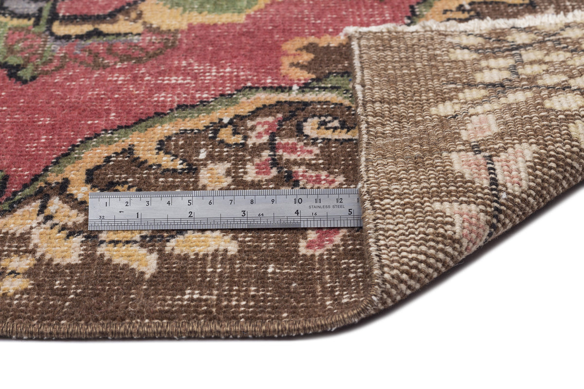 Red, brown, and green floral medallion rug with a folded corner showing the woven backing and a metal ruler for scale.