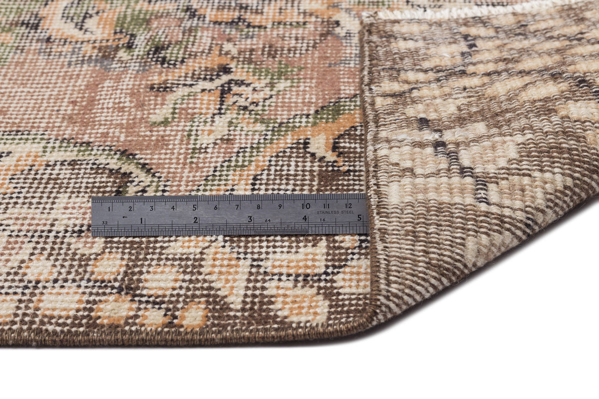 Distressed floral rug in brown, tan, and green with a folded corner revealing the backing and a metal ruler for scale.