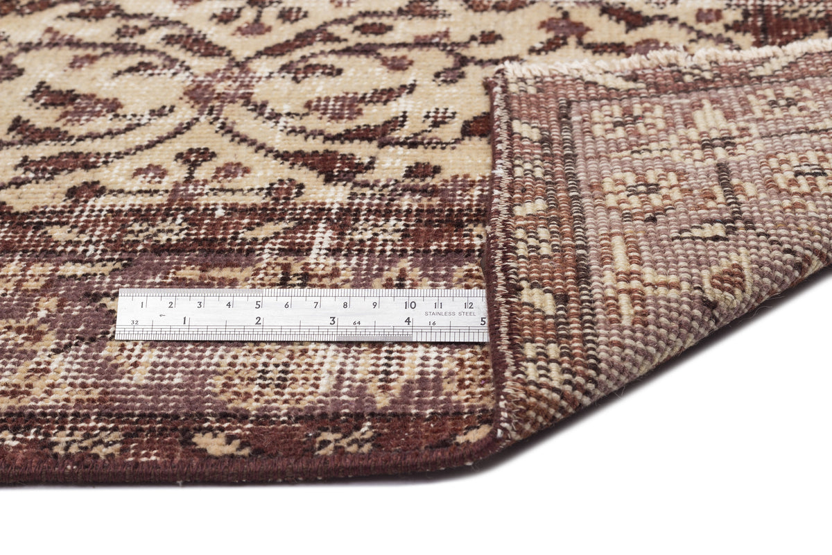 Beige and brown traditional floral rug with a folded corner showing the woven backing and a metal ruler for scale.