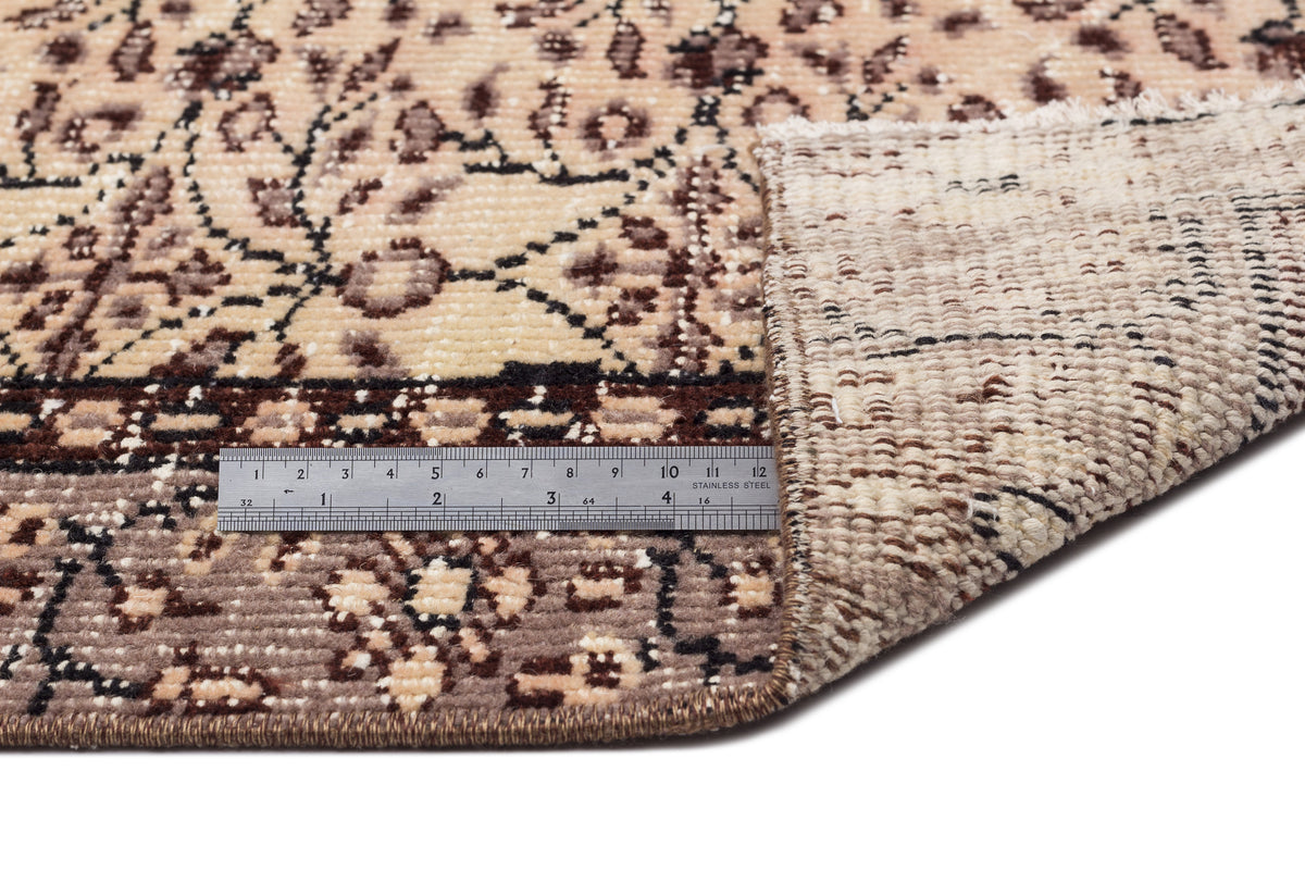 Beige and brown floral patterned rug with a folded corner revealing the backing and a metal ruler for scale.