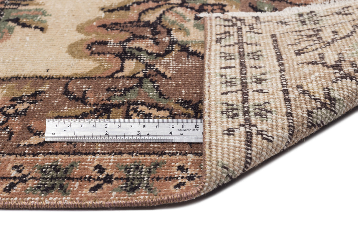 Beige, brown, and green traditional floral rug with a folded corner showing the woven backing and a metal ruler.