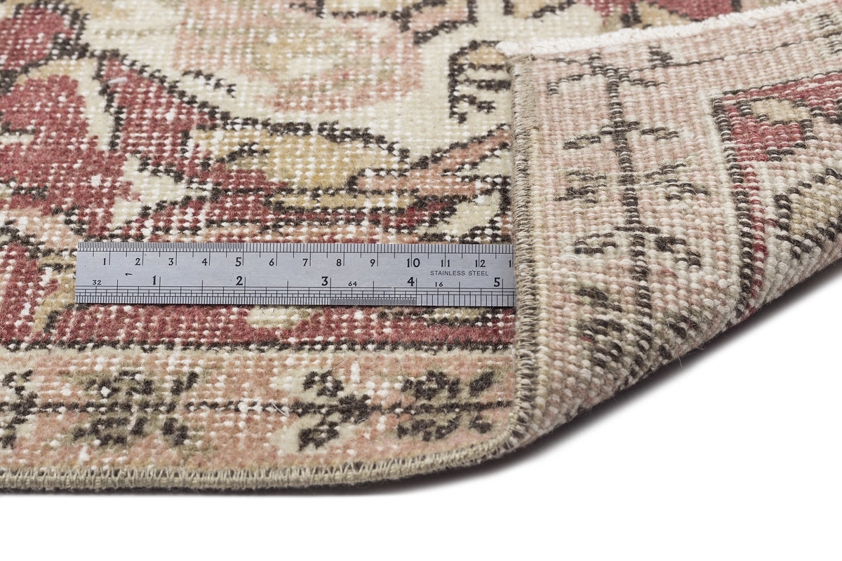 Distressed vintage-style rug in cream, red, and brown with a floral medallion pattern, shown with a metal ruler for scale.