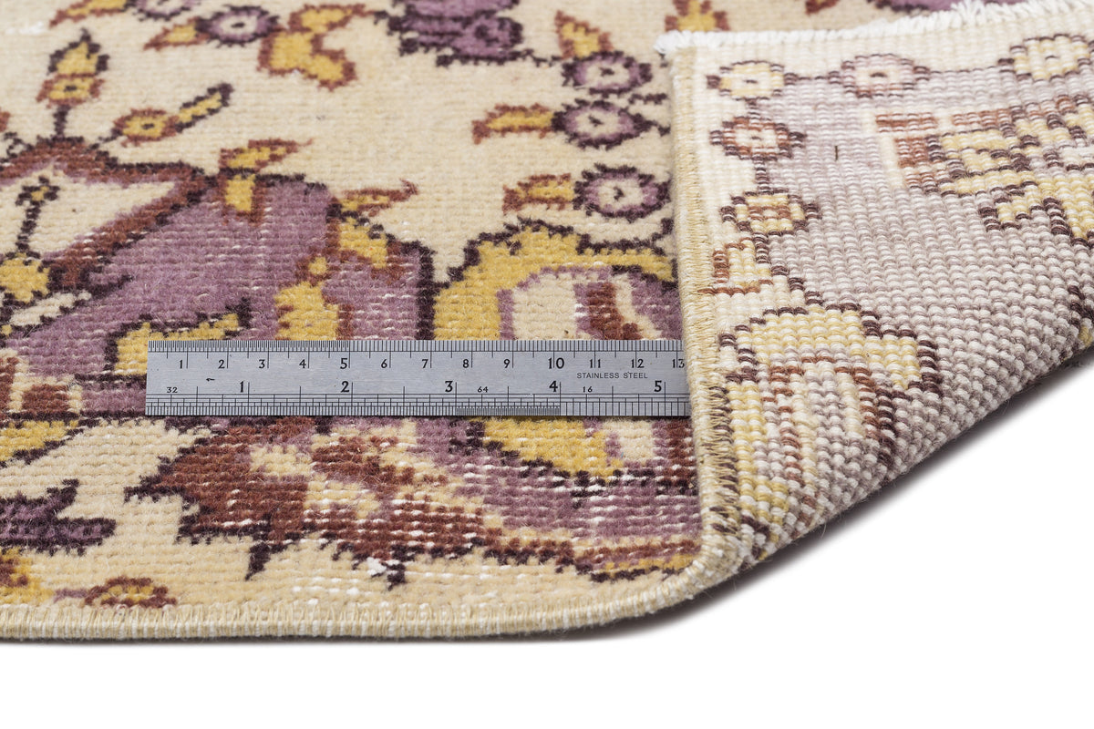 Beige rug with a purple and yellow floral pattern, shown folded to reveal the woven backing and a metal ruler for scale.