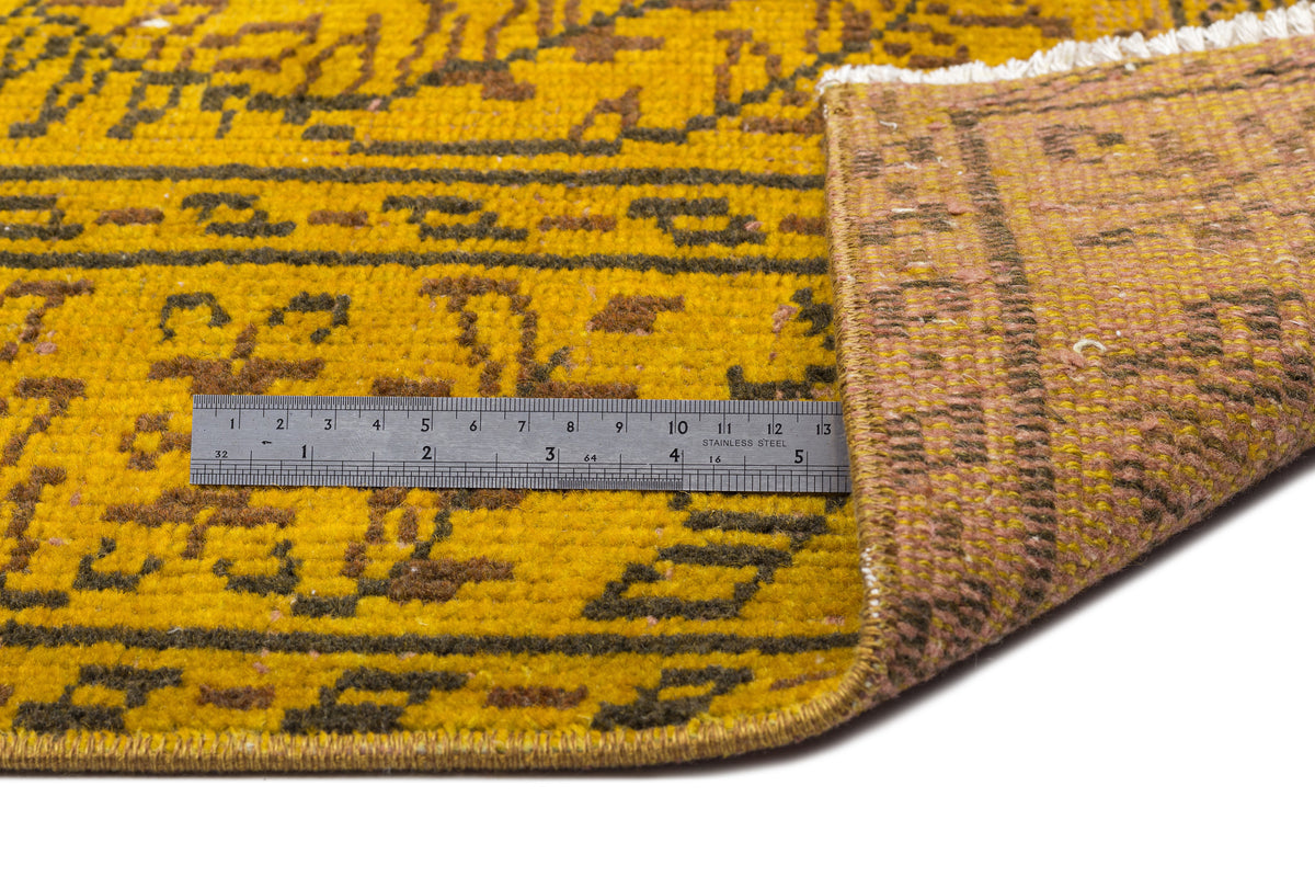 Mustard yellow and brown patterned rug with a folded corner revealing the backing, shown with a metal ruler for scale.