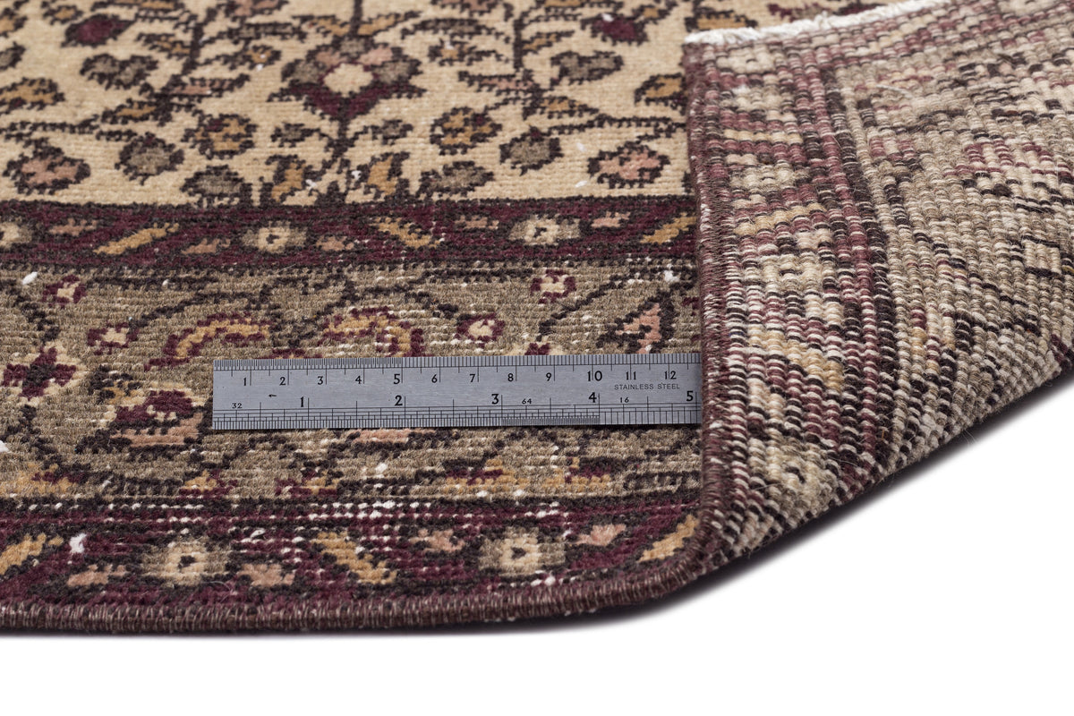 Beige and burgundy traditional floral rug with a folded corner revealing the woven backing and a metal ruler for scale.
