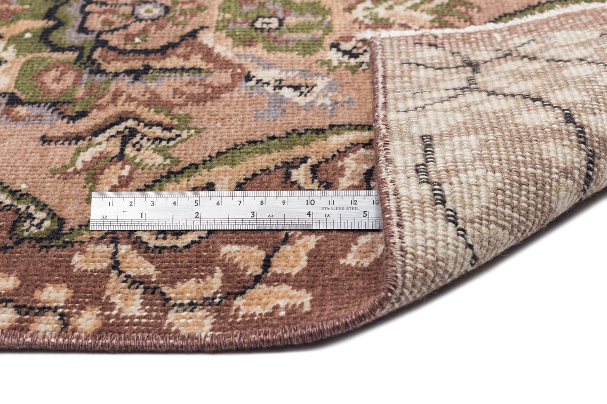 Brown, green, and beige floral medallion rug with a folded corner revealing the woven backing and a metal ruler.