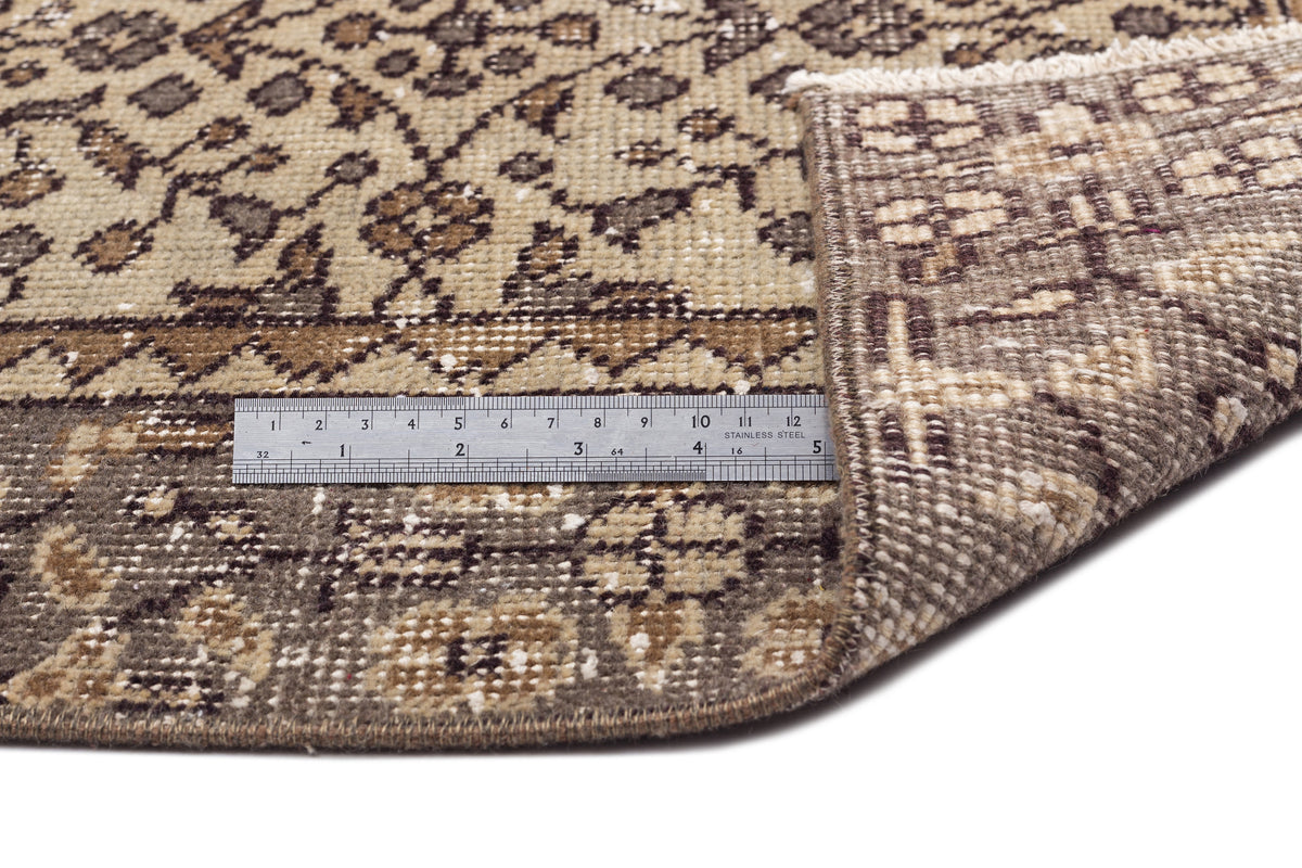 Brown and beige traditional patterned rug with a folded corner and a metal ruler for scale on a white background.