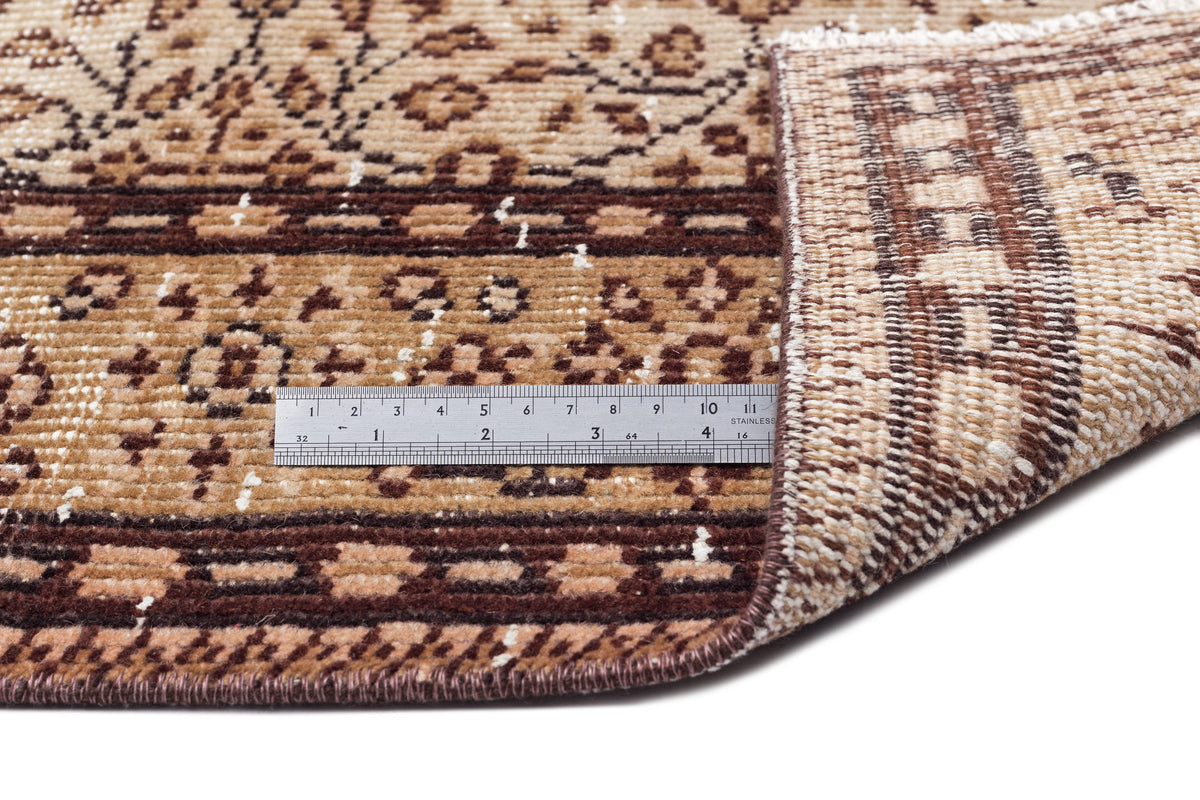 Brown and tan patterned rug with a folded corner showing the weave and a metal ruler for scale.