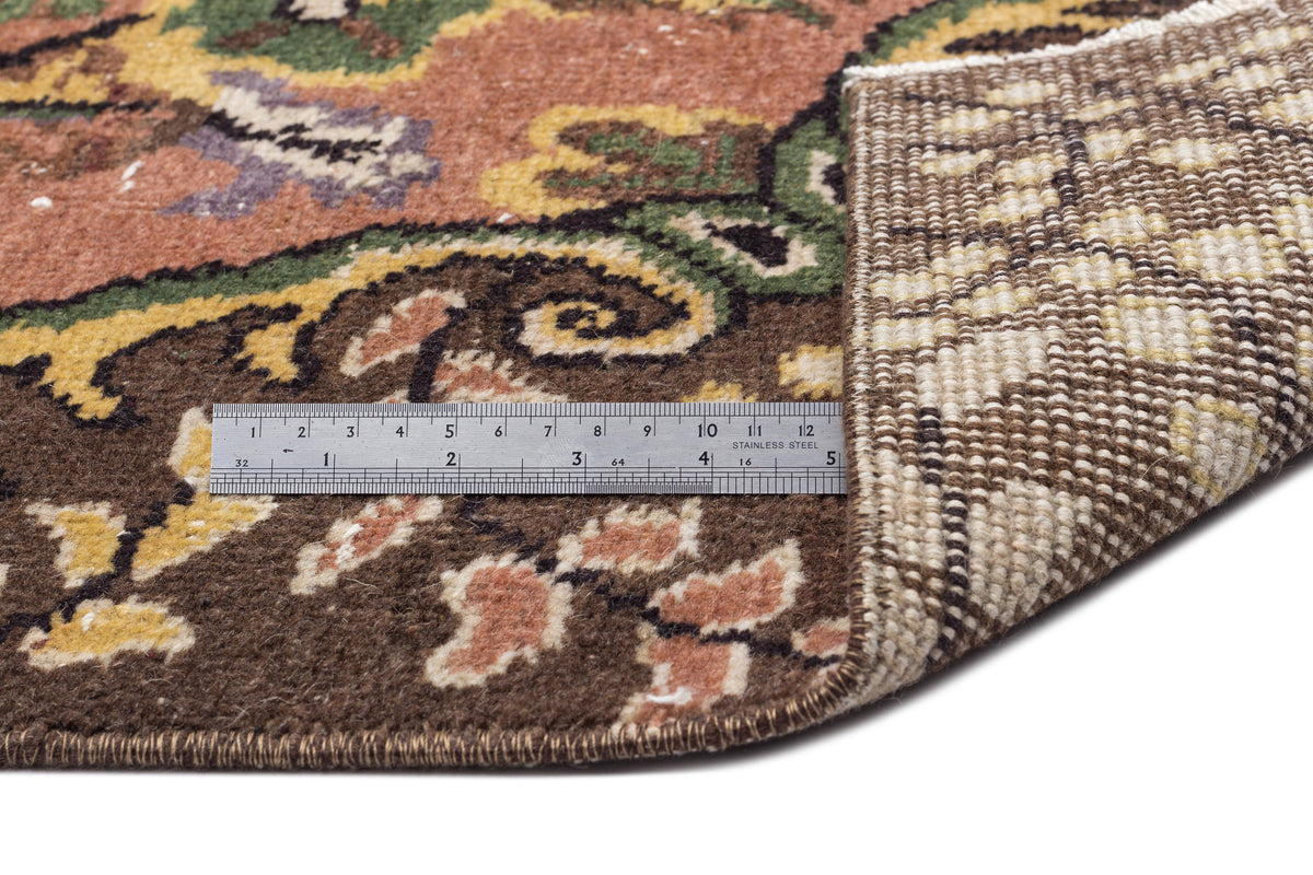 Floral patterned rug in brown, green, and yellow with a folded corner showing the woven backing and a metal ruler.