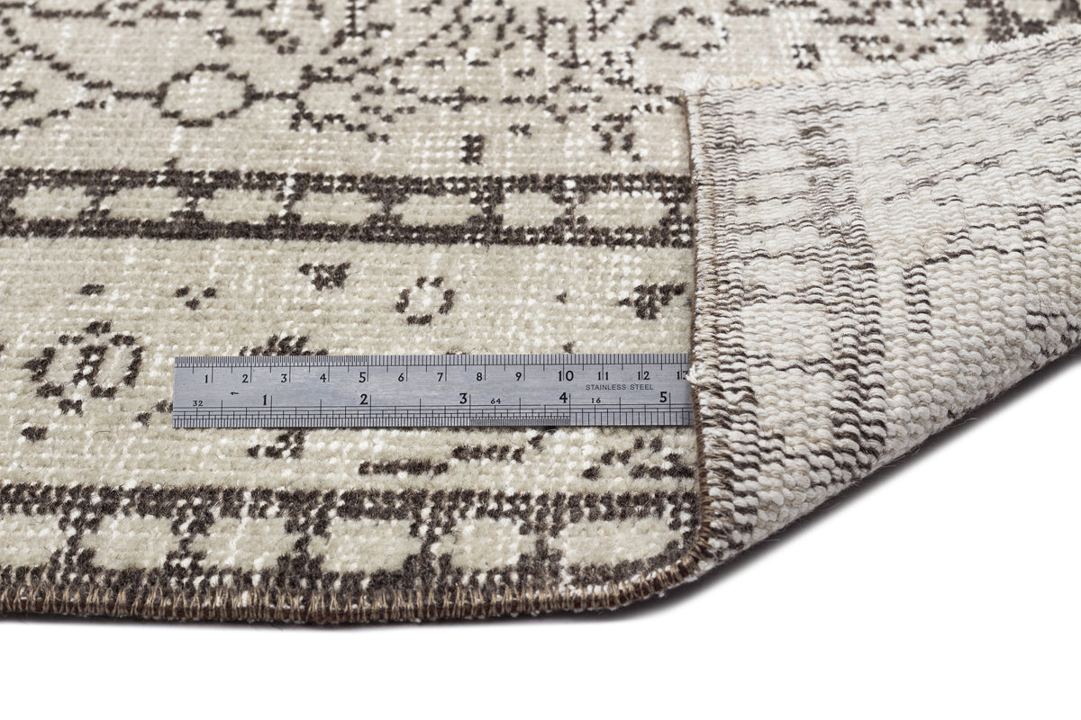 Beige and dark brown patterned rug with a folded corner showing the backing, measured by a stainless steel ruler.