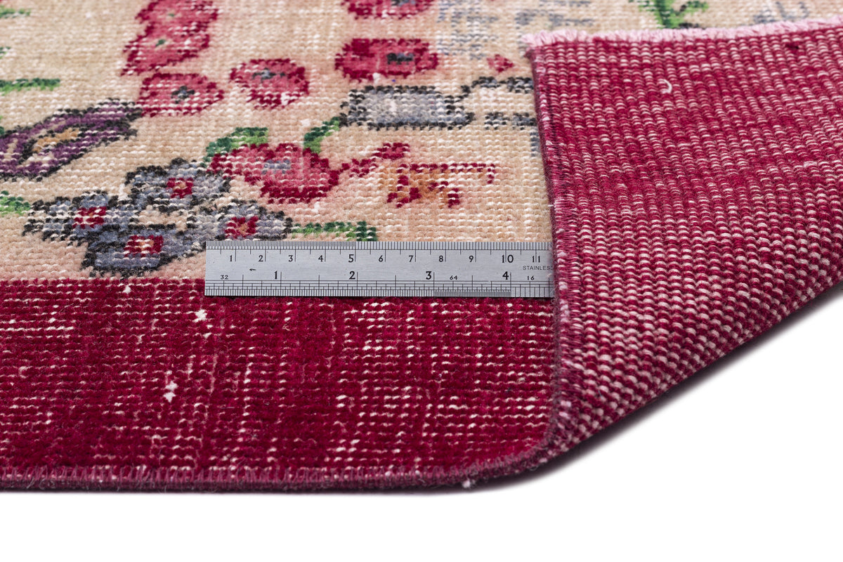 Beige rug with a floral pattern in red, purple, and green, featuring a deep red border and a metal ruler for scale.