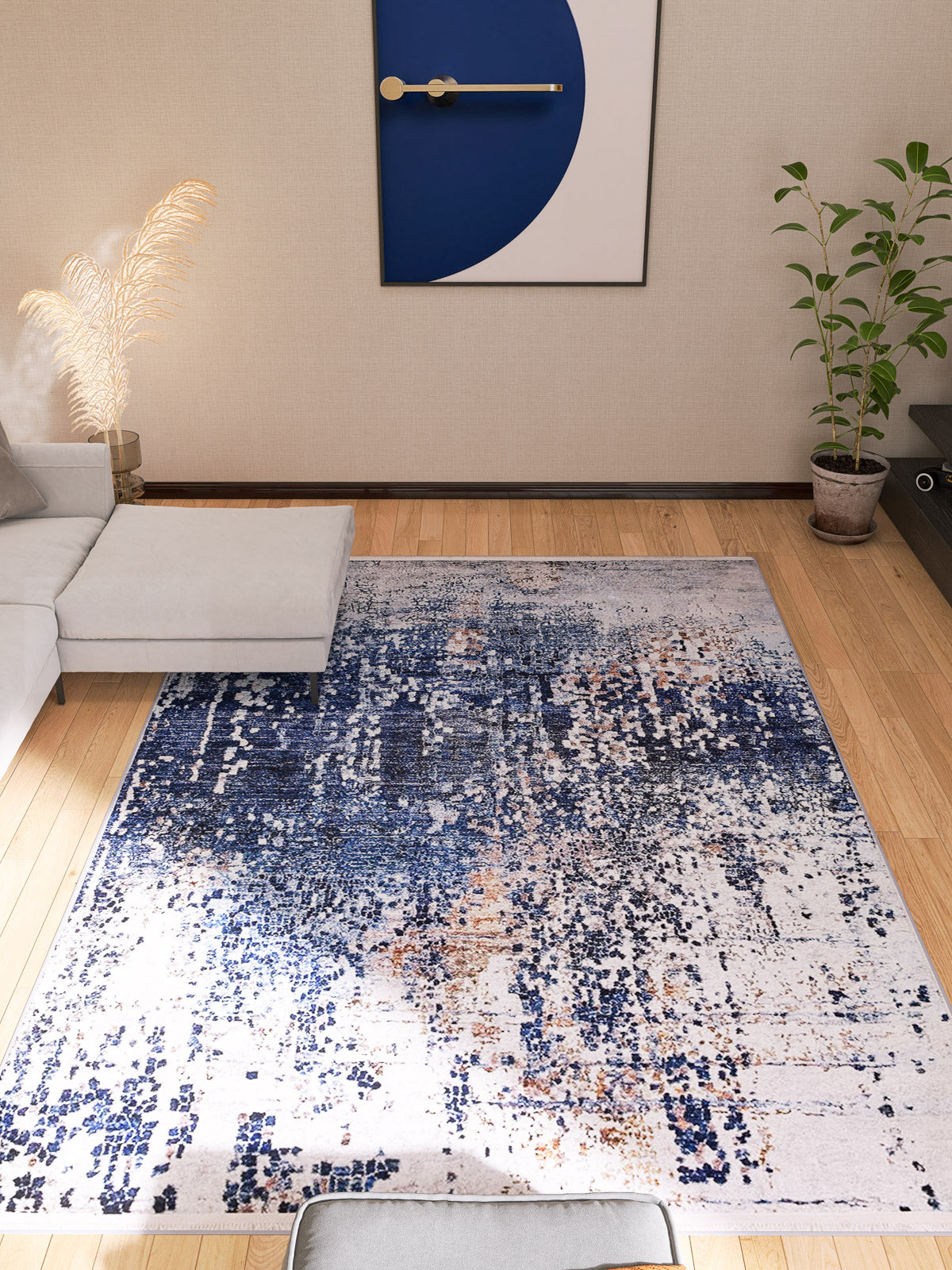 A modern living room with a light gray sectional sofa, potted plants, and the Abstract Atmosphere - Washable Rug - JR834 in blue and white as both a flat weave rug and large geometric wall art above the sofa on a light wall.