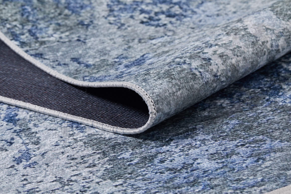 A close-up of the Abstract Oasis - Washable Rug - JR144 (Custom Size) in blue and gray shows its textured abstract pattern. One corner is lifted, revealing the dark backing and stitched edge of this allergy-friendly rug.