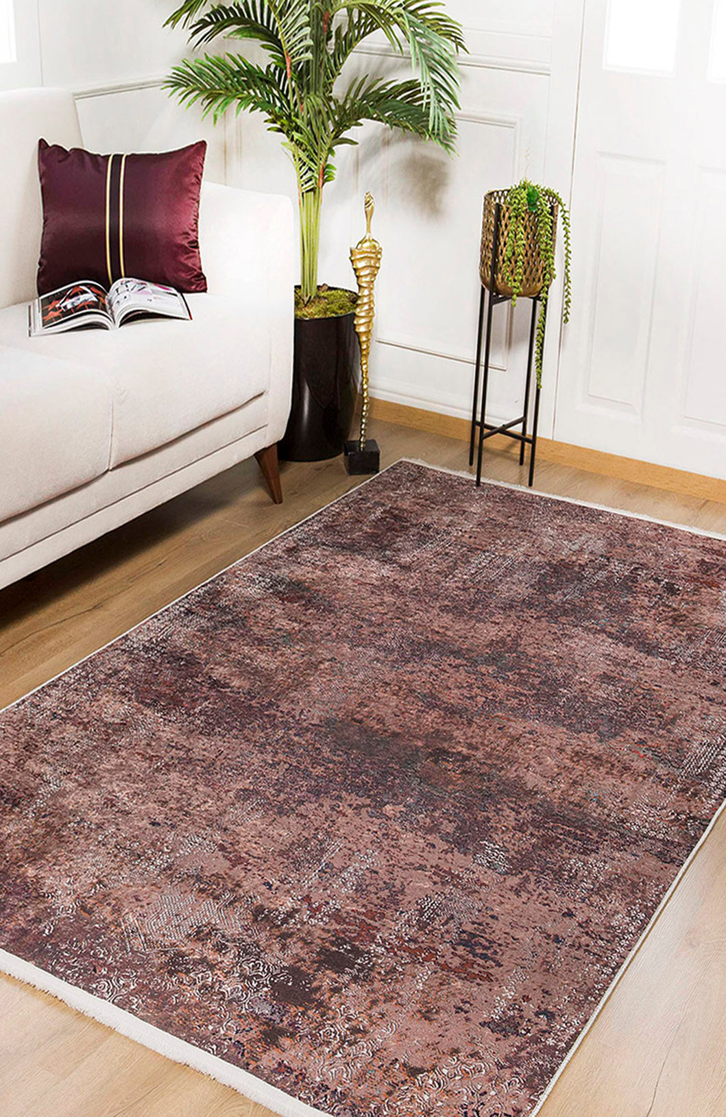 A living room featuring the Abstract Spectrum - Washable Rug - JR1427 (Custom Size) in bronze and brown on a wooden floor, a beige sofa with a burgundy pillow, an open book, tall green plant, small statue, and potted plant on black stand.