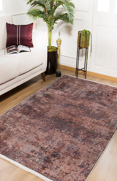 Load image 1 in gallery view. A living room featuring the Abstract Spectrum - Washable Rug - JR1427 (Custom Size) in bronze and brown on a wooden floor, a beige sofa with a burgundy pillow, an open book, tall green plant, small statue, and potted plant on black stand.