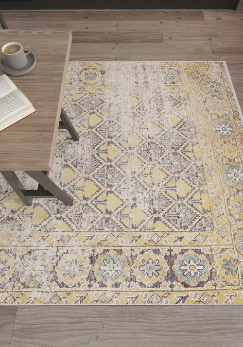 Bild 1 in Galerieansicht laden. The Aged Splendor - Washable Rug - JR1906 (Custom Size), featuring yellow-gold with gray and white geometric and floral designs, lies on a wooden floor beside a table with coffee and an open book—ideal for high-traffic areas.
