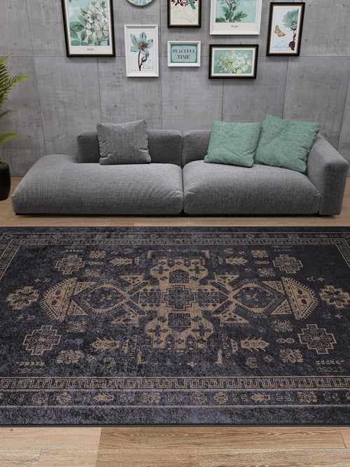Load image 1 in gallery view. A modern living room features a gray sectional, green and gray pillows, framed botanical art on a concrete wall, and the Ancient Legacy - Washable Rug - JR1890 (Custom Size) in black. A potted plant sits to the left on wooden flooring.