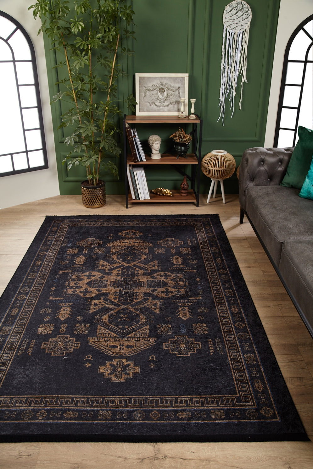 A stylish living room with an Ancient Legacy - Washable Rug - JR1890 (Custom Size) in black and gold, a gray tufted sofa with green pillows, shelves of books and decor, a tall plant, and green walls with framed art and woven accents.
