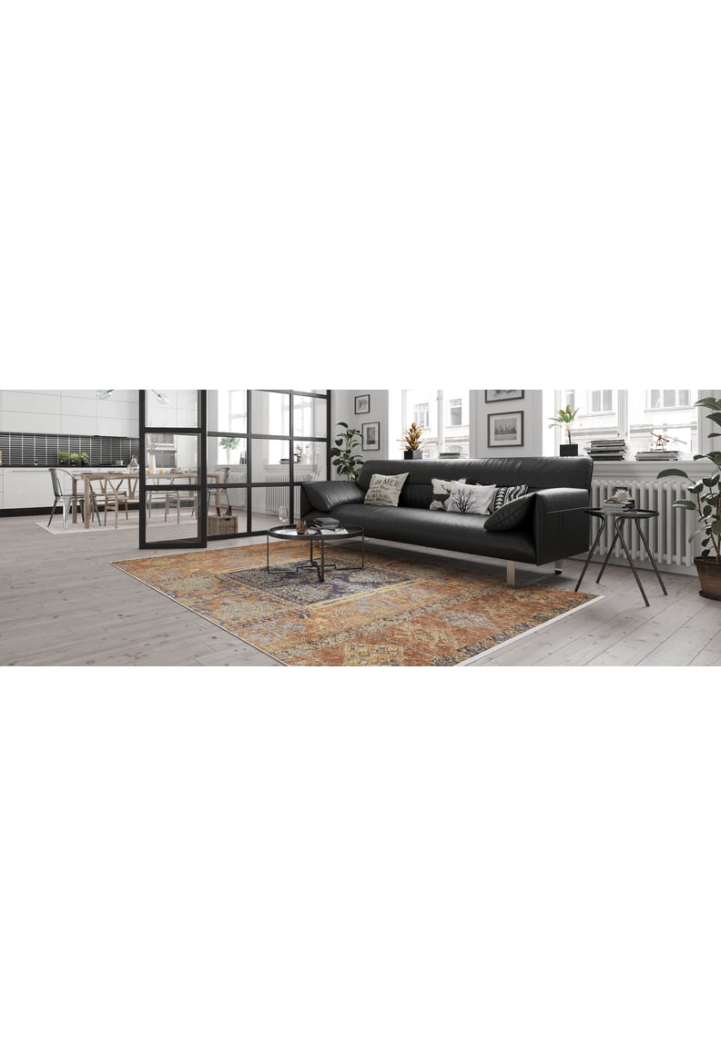 A modern living room features a black sofa, coffee table, throw pillows, potted plants, and the Ancient Palace - Washable Rug - JR1864 in vibrant orange. Large windows and glass doors reveal a kitchen with light wood floors ideal for high-traffic areas.