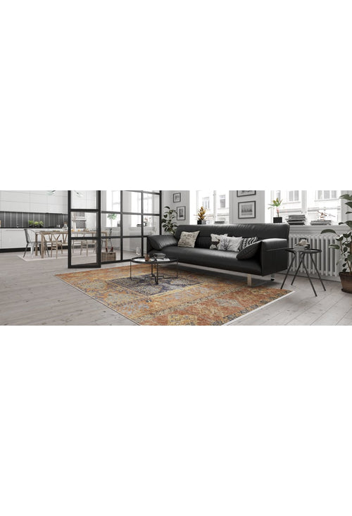Load image 1 in gallery view. A modern living room features a black sofa, patterned pillows, and the Ancient Palace - Washable Rug - JR1864 (Custom Size) in orange—ideal for high-traffic areas. A small coffee table, large windows, plants, plus a dining area and white-cabinet kitchen complete the space.