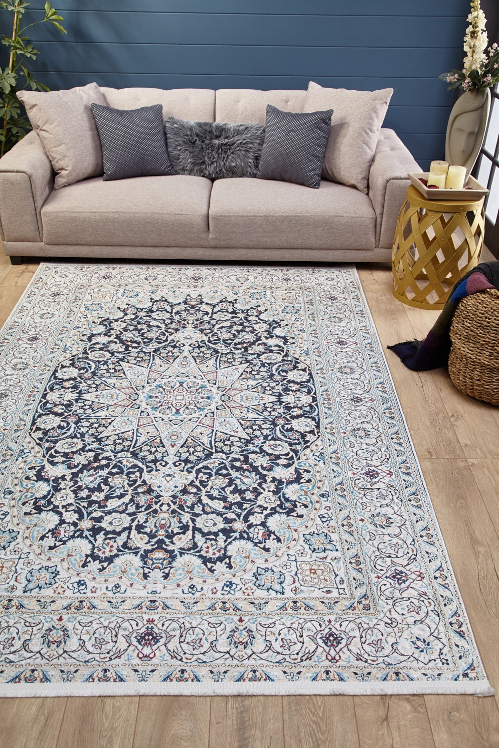 A living room with a beige sofa, gray furry pillows, and the Antique Enchantment - Washable Rug - JR1686 (Custom Size) in blue and white on wooden floors. A round yellow side table with candles and a woven basket holding a blanket sit beside the sofa.