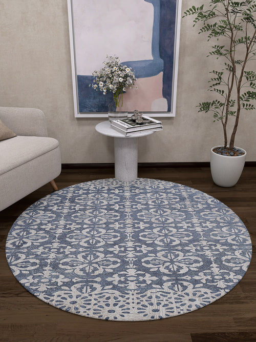Load image 1 in gallery view. The Antique Flourish - Washable Rug - JR1591 (Custom Size) features a round blue and white pattern, ideal for cozy living rooms or high-traffic areas, shown on a dark wooden floor beside a light sofa, side table with books, plant, and abstract wall art.