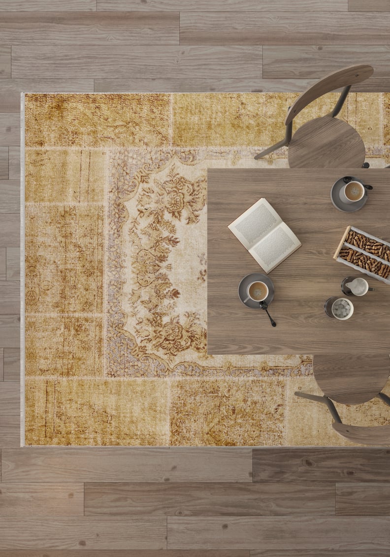Top-down view of a wooden table with two chairs, coffee cups, an open book, and snacks atop the Antique Insignia - Washable Rug - JR309 in yellow-gold tones over a wooden floor.