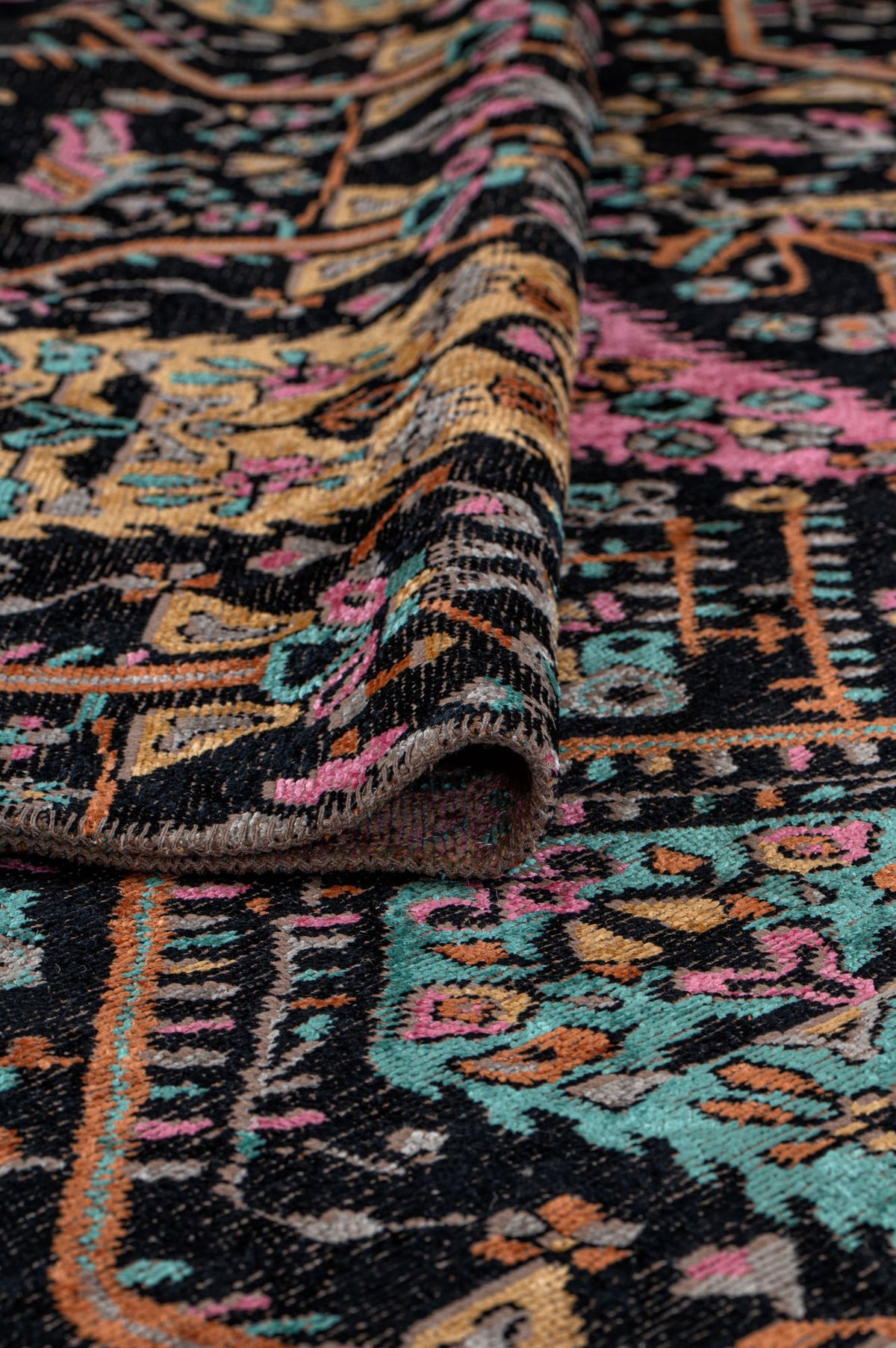 A close-up of the Midnight Bazaar Area Rug APX1911, featuring colorful flatwoven geometric and floral patterns in pink, yellow, teal, and black. The rug is slightly folded to highlight its texture and thickness.