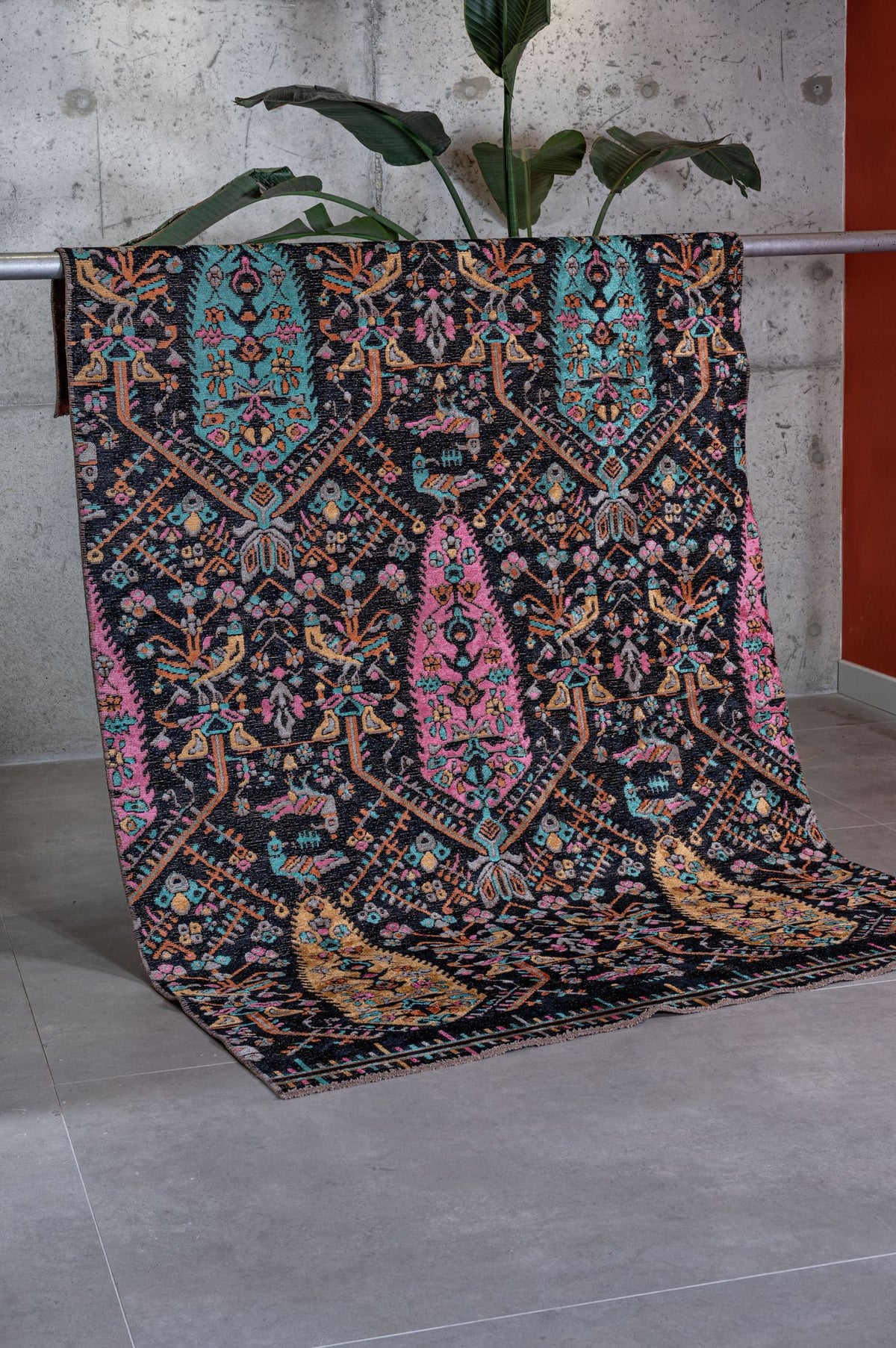 The Midnight Bazaar Area Rug APX1911, featuring intricate flatwoven designs in turquoise, pink, yellow, and white, is draped over a metal rail by a gray concrete wall with a green plant in the background.