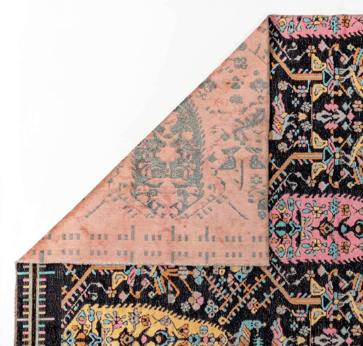 A corner of the Midnight Bazaar Area Rug APX1911 is flipped over, showing its muted pink reverse side. The rug’s front displays intricate, colorful floral and geometric patterns on a dark background.