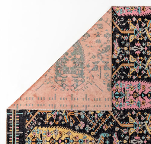 Bild 1 in Galerieansicht laden. A corner of the Midnight Bazaar Area Rug APX1911 is flipped over, showing its muted pink reverse side. The rug’s front displays intricate, colorful floral and geometric patterns on a dark background.