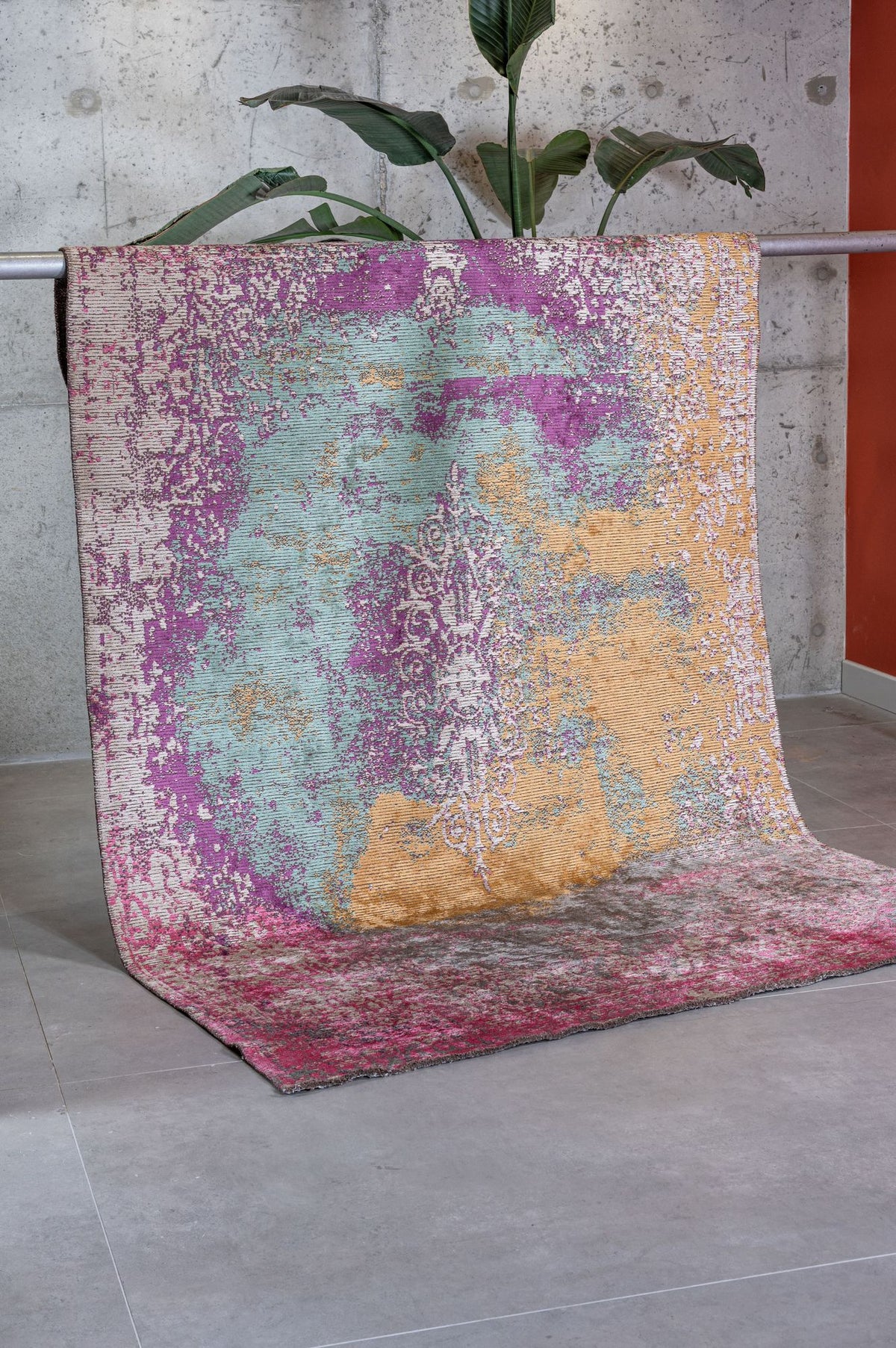 Distressed abstract rug in magenta, teal, and gold with a faded central medallion, draped against a concrete wall.