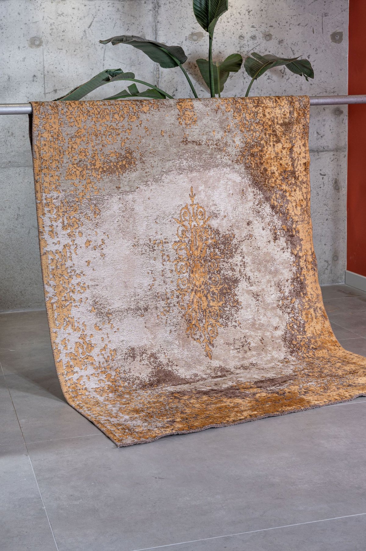 The Golden Fade Area Rug APX1922, featuring a gold and beige pattern, is draped over a metal railing before a gray concrete wall. A lush green plant peeks from behind, with light gray tile flooring underneath.