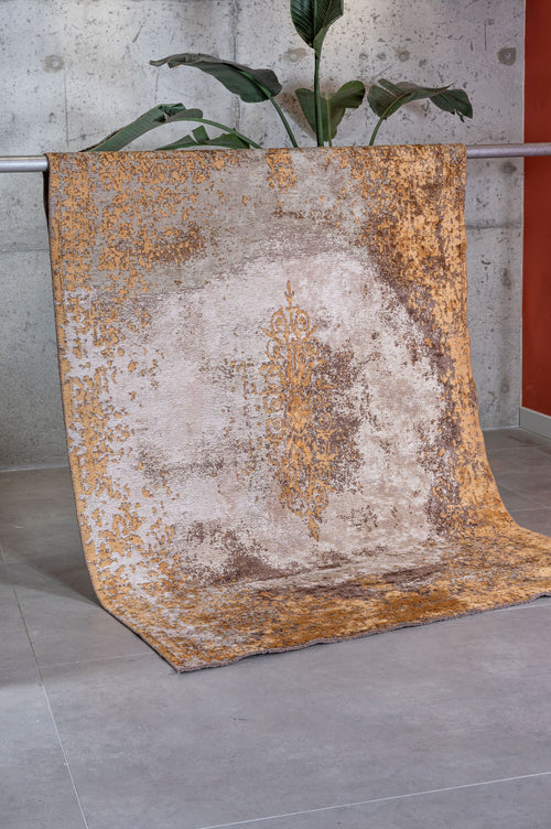 Load image 1 in gallery view. The Golden Fade Area Rug APX1922, featuring a gold and beige pattern, is draped over a metal railing before a gray concrete wall. A lush green plant peeks from behind, with light gray tile flooring underneath.
