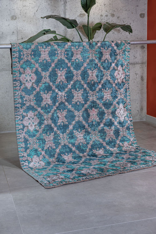 Load image 1 in gallery view. The Ocean Trellis Area Rug APX1942, featuring blue and pink hues in a modern flatwoven design, is draped over a metal bar in a contemporary room with a concrete wall, gray tile floor, and a tall green plant.
