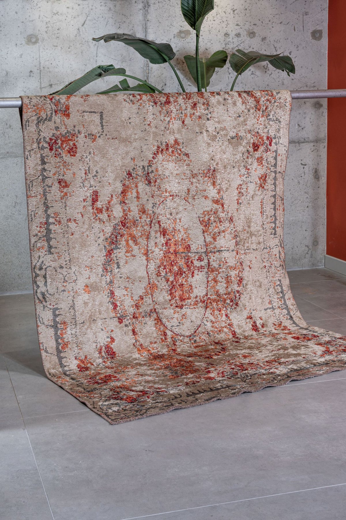 Beige and rust-red distressed medallion rug draped over a metal bar against a concrete wall.