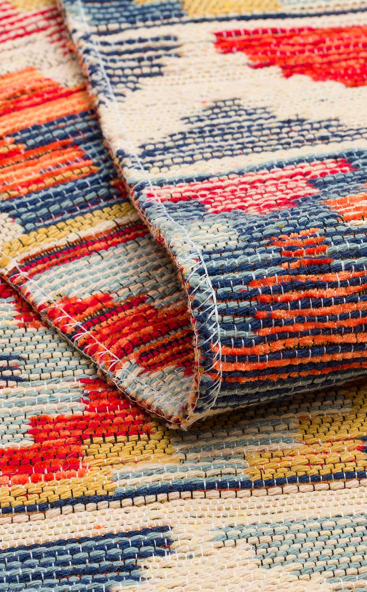 A detailed view of the Aqua Flow - Washable Reversible Kilim - NMD02 shows its vibrant red, blue, yellow, and cream patterns. The folded corner highlights the textured weave and striking geometric designs of this unique area rug.