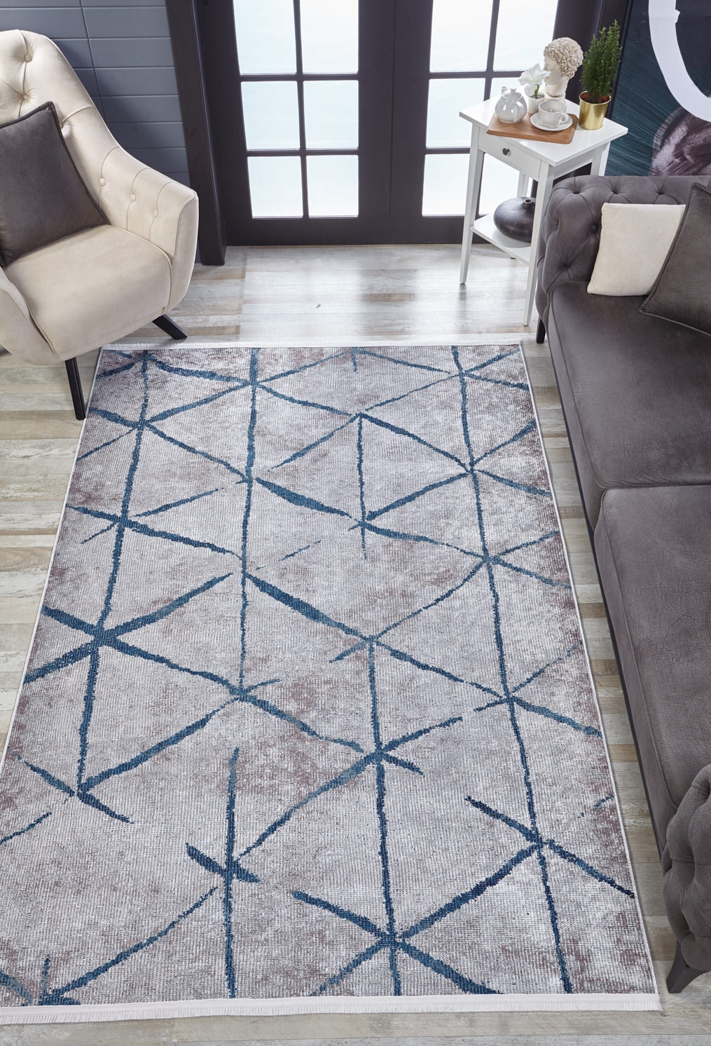 A modern living room features a beige tufted armchair, gray sofa, white side table, and the Arabesque Trellis - Washable Rug - JR1732 (Custom Size) with blue geometric lines—ideal for allergy-sensitive homes. Large glass doors complete the scene.