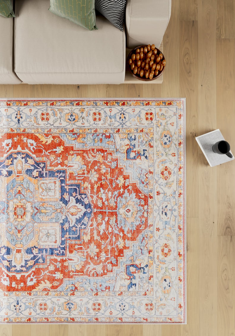 A beige sofa with striped cushions sits next to the Ardentis Medallion Washable Rug - LCC3002 (Outlet) US on a light wooden floor, with a bowl of snacks and a magazine beside a mug nearby.