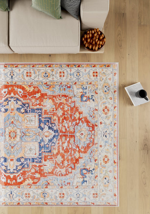 Load image 1 in gallery view. A top-down view of the Ardentis Medallion Washable Rug - LCC3002 (Custom Size) in red multicolor with blue and beige designs, placed next to a beige sofa, a bowl of dates, and a mug on an open book on a light wooden floor.