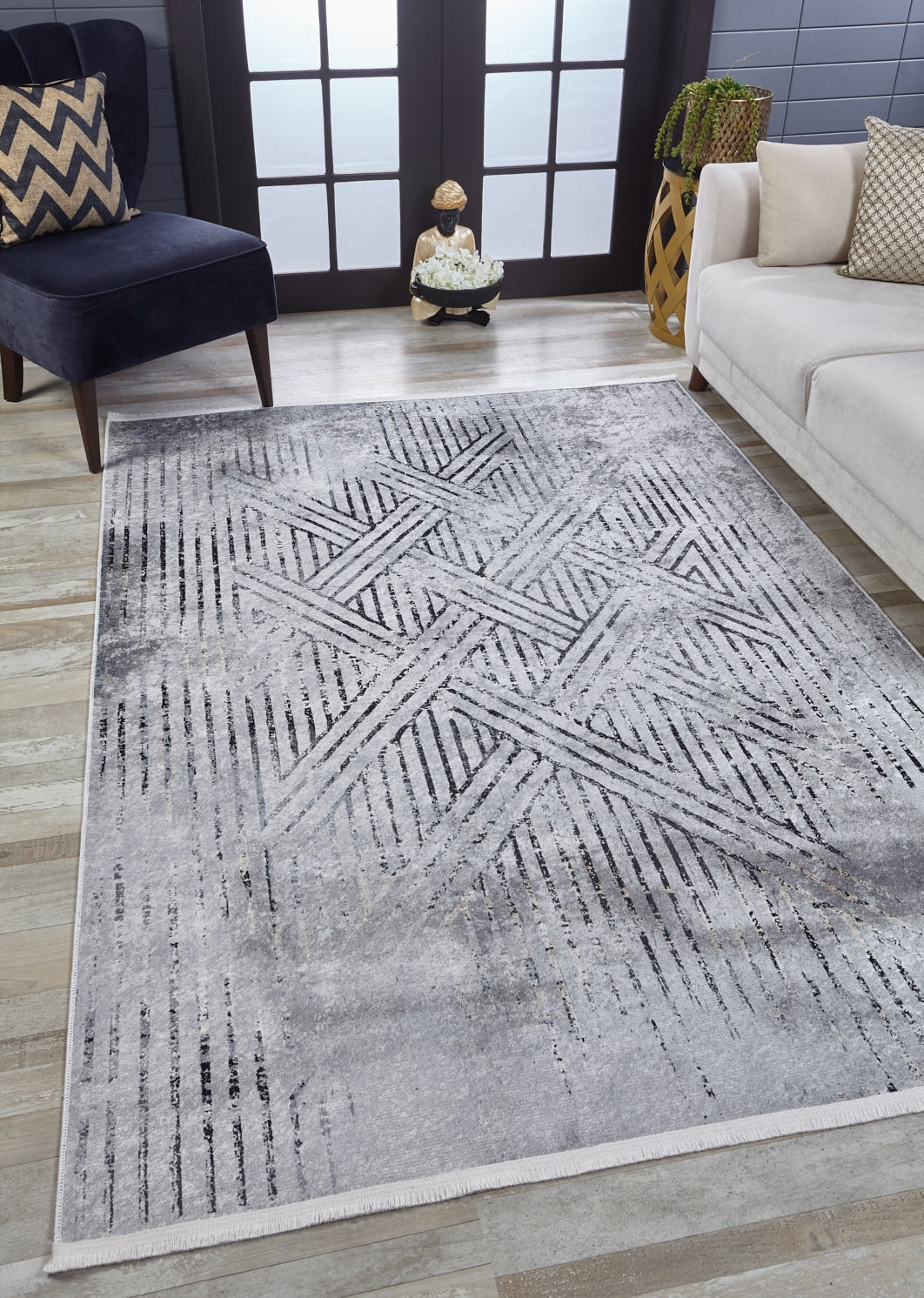 A living room features the Art Deco Empress - Washable Rug - JR1867 (Custom Size), a modern light gray-anthracite geometric rug, paired with a white sofa, dark blue chevron pillow chair, decorative basket, and large black-framed windows.