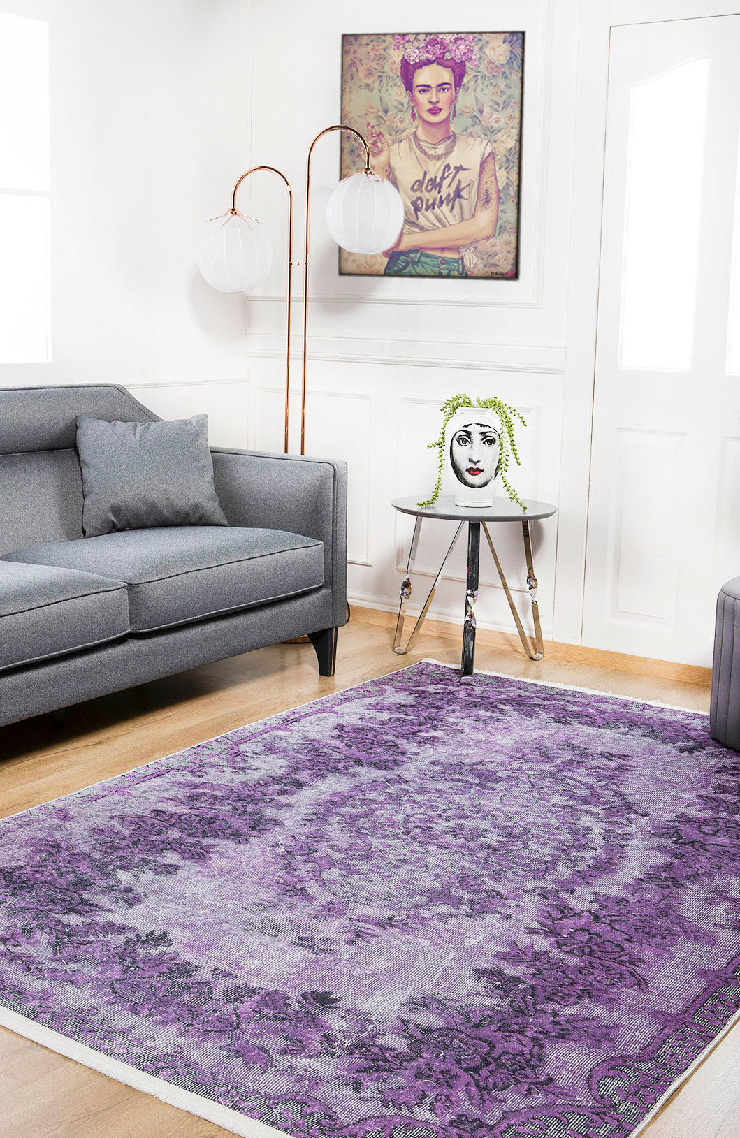 A modern living room with a gray sofa, an Artisan Tapestry - Washable Rug - JR176 (Custom Size) in purple for high-traffic areas, a round side table with a face vase, two floor lamps, and a colorful framed portrait on the wall.