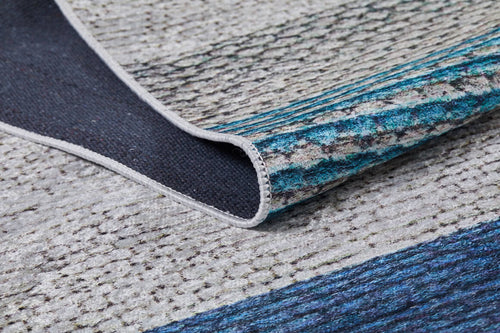 Load image 1 in gallery view. A close-up of the Artistic Angles - Washable Rug - JR908 (Custom Size), a flat-weave rug in blue, teal, and grey tones, showing its folded edge to reveal the dark backing and stitched border.