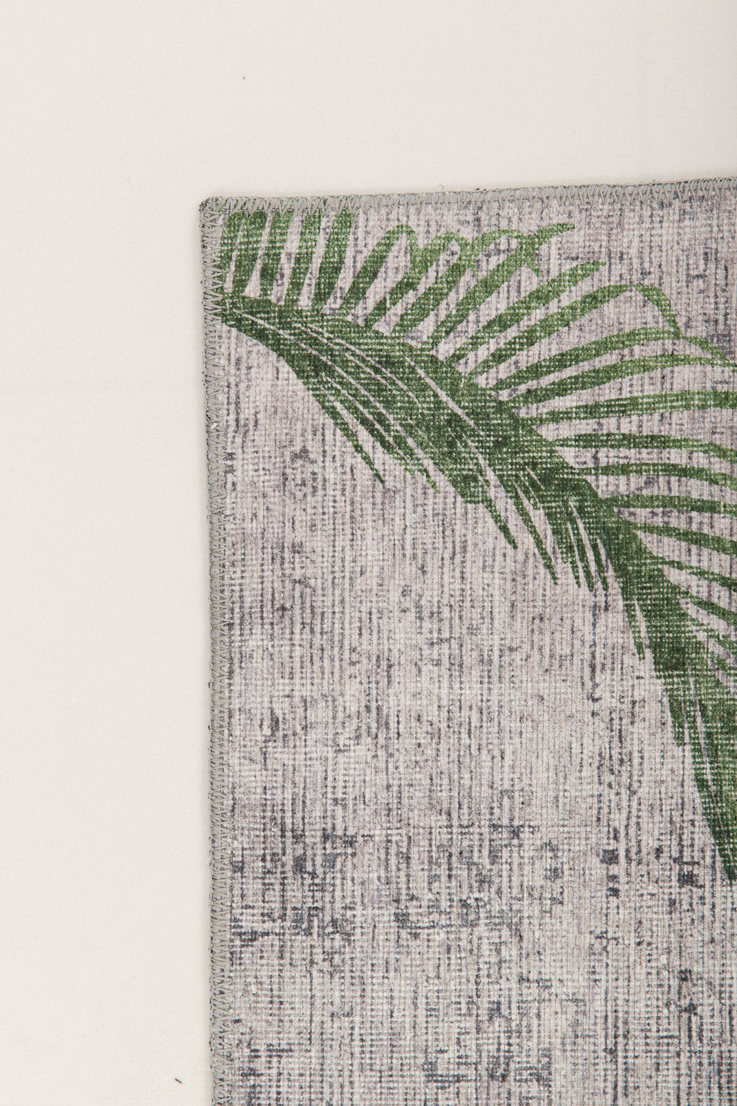 A green palm leaf print accents the upper left corner of the Ashwing Silence - Washable Rug - JR0048, featuring a textured, light-colored surface set against a plain light background.