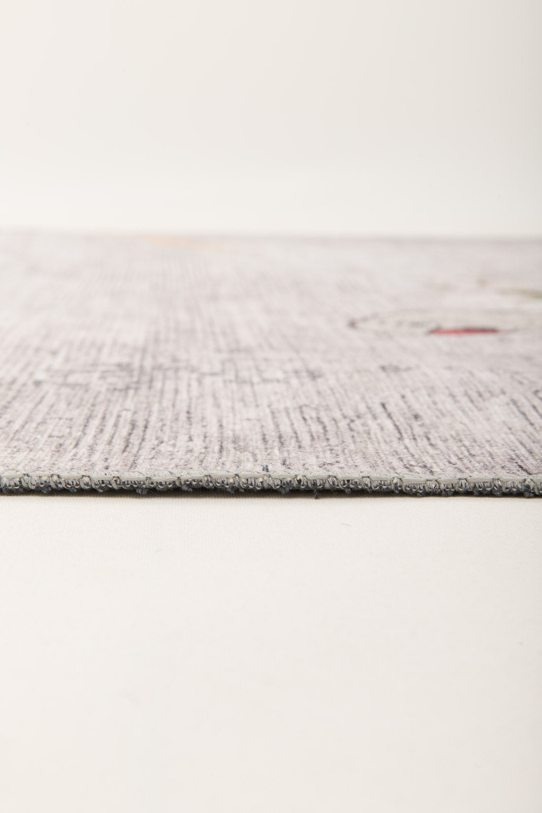 Close-up side view of the Ashwing Silence - Washable Rug - JR0048, featuring a thin, light-colored textured surface with subtle patterns on a neutral background. The edge is in focus, showcasing its low-profile design.