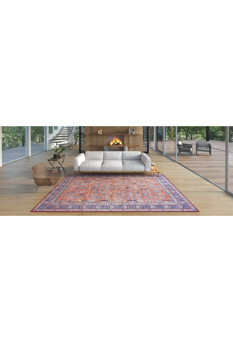 A modern living room with a white sofa, wood-paneled fireplace, Aurelius Coral Washable Rug - LCC3005 featuring colorful patterns, a wicker chair, and large glass windows overlooking greenery and a patio—ideal for high-traffic areas.