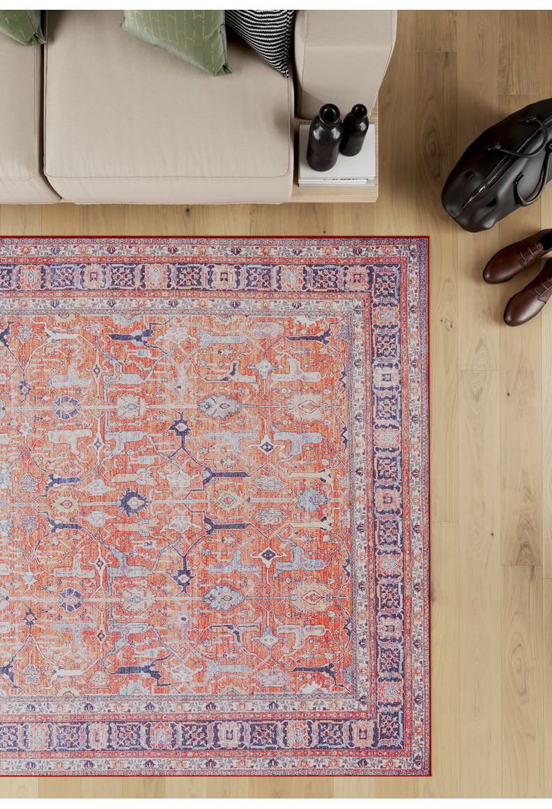 A beige sofa with green and striped pillows sits beside the Aurelius Coral Washable Rug - LCC3005 on a wooden floor. Nearby are a black bag, brown shoes, and two dark bottles on a small white side table.
