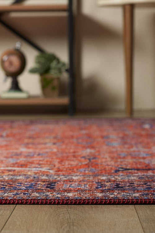 Load image 1 in gallery view. A close-up of the Aurelius Coral Washable Rug - LCC3005, featuring red and purple patterns on a wooden floor, perfect for high-traffic areas, with blurred shelves, a plant, and decor in the background.