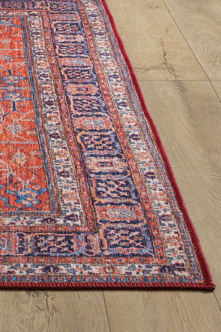 A close-up of the Aurelius Coral Washable Rug - LCC3005 shows its colorful, patterned flat weave in red, purple, and orange on a light wood floor. The detailed border and visible woven texture offer both style and durability for high-traffic areas.